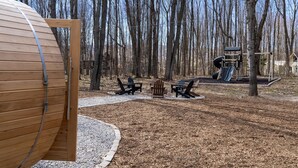 A wooden cabin with a door open, surrounded by a forest. There are Adirondack chairs and a playground with slides. - Built for You - 4BR w/ Playground, Sauna, Games! (Harbor Springs)