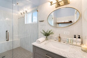 A modern bathroom with a glass-enclosed shower, a marble sink, a large mirror, and toiletries on the countertop.