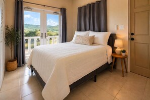 Une chambre à coucher avec un lit, des rideaux, une table de chevet avec une lampe, une plante en pot et une vue sur la verdure à l’extérieur.