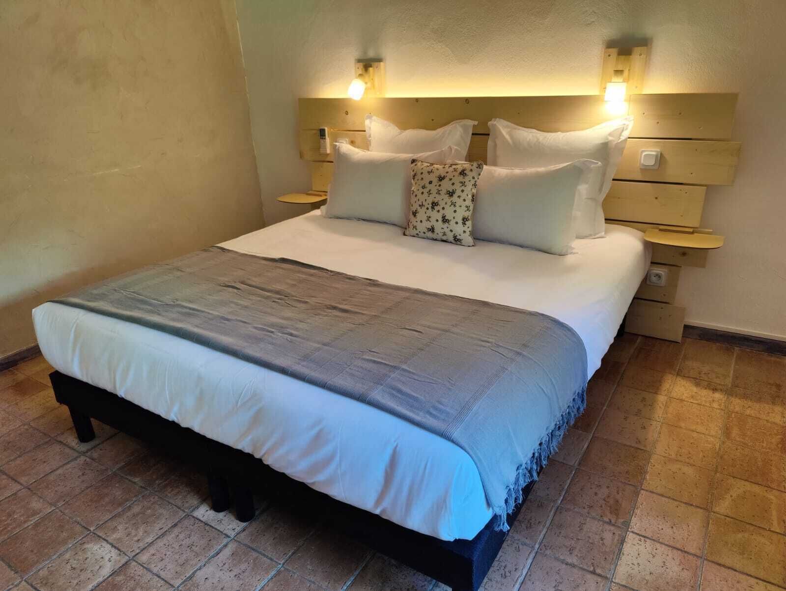 Una cama bien hecha con una manta gris, almohadas blancas y un cabecero de madera con luces fijadas a la pared.