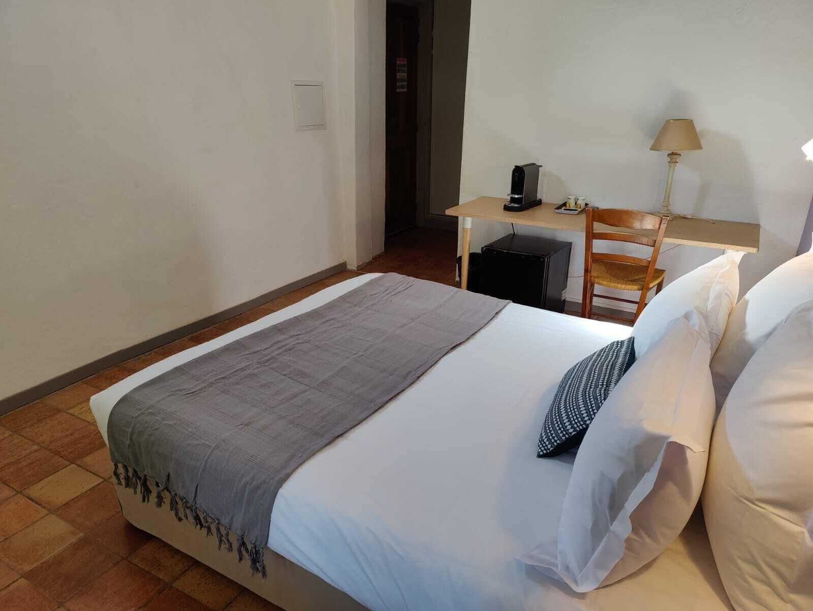 Un lit simple avec une couverture grise et des oreillers blancs, un bureau avec une lampe et une chaise, dans une pièce aux murs blancs et au sol carrelé.