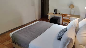 Un lit simple avec une couverture grise et des oreillers blancs, un bureau avec une lampe et une chaise, dans une pièce aux murs blancs et au sol carrelé.