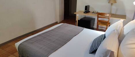 A single bed with a grey blanket and white pillows, a desk with a lamp, and a chair in a room with white walls and a tiled floor.
