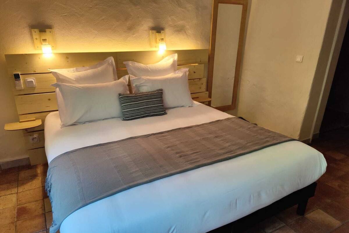 Cama bem arrumada com lençóis brancos e manta cinza, cabeceira de madeira com luminárias fixadas na parede e mesa de cabeceira de madeira com uma gavetinha.