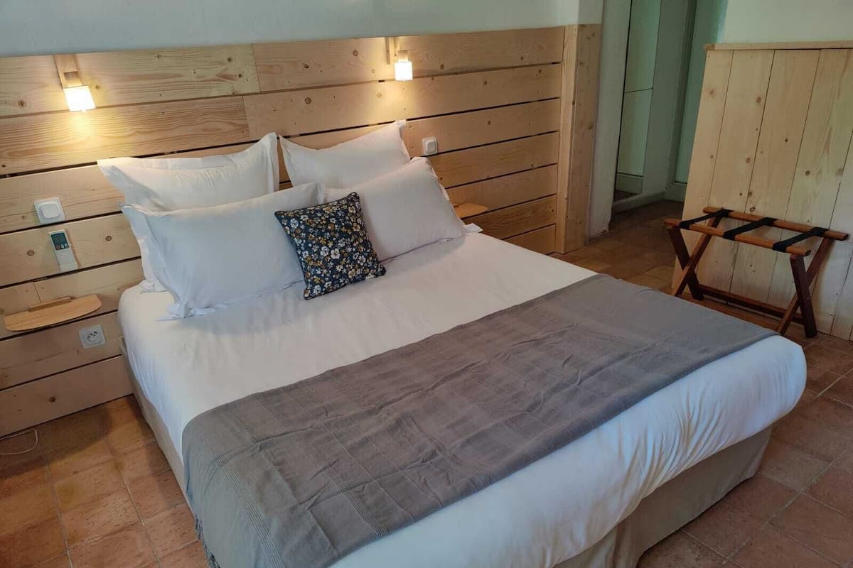 Quarto com cabeceira de madeira, cama com roupa de cama branca e cinza, mesinha de cabeceira e um banco de madeira.