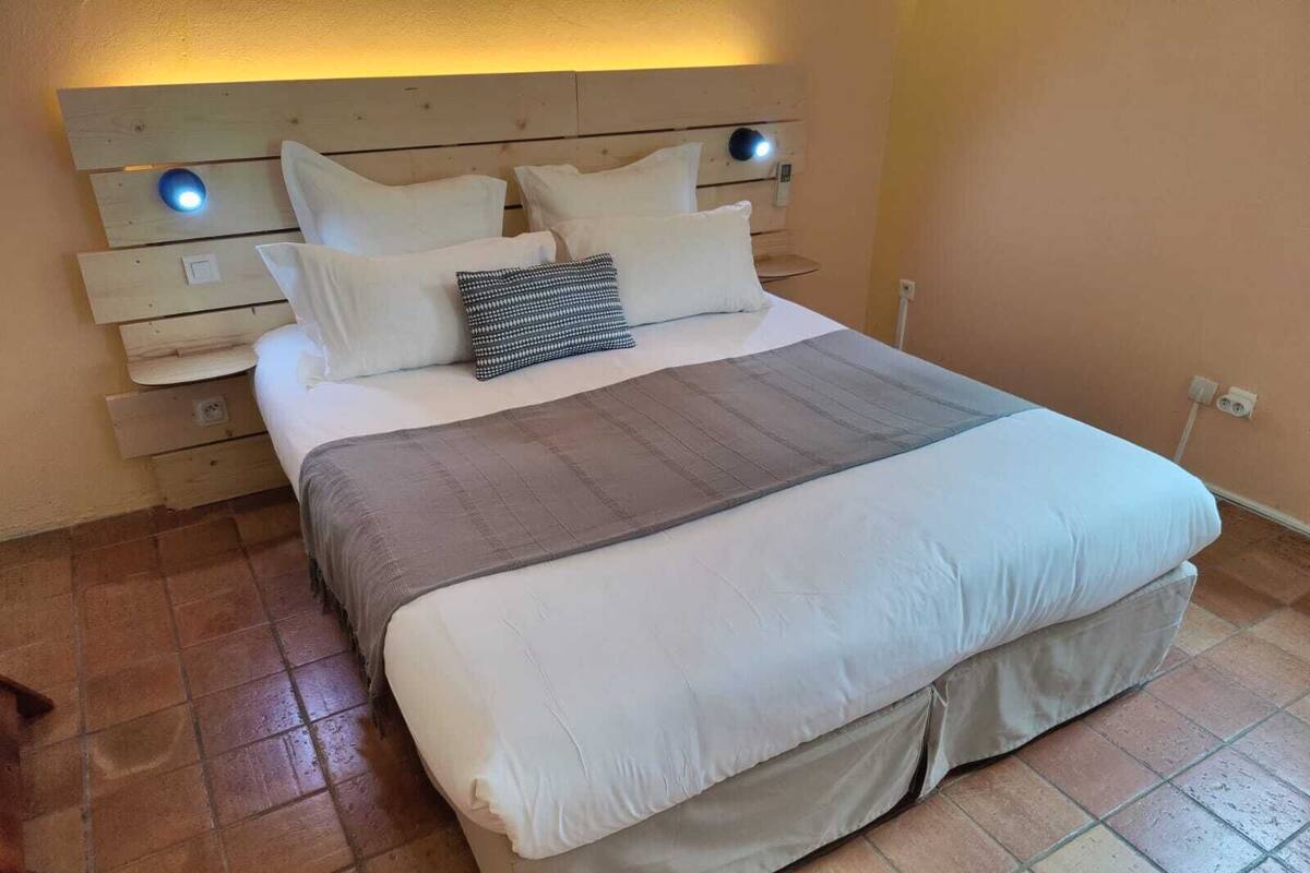 Cama bem arrumada com manta cinza e branca listrada, almofadas brancas e cabeceira com duas luzes fixadas na parede.