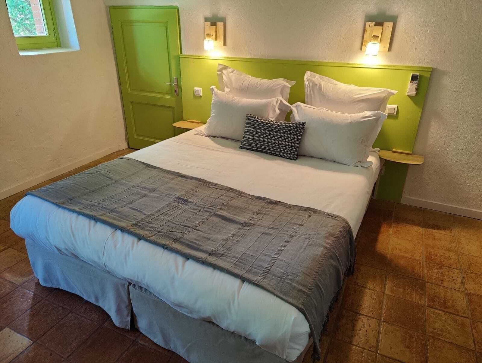 A neatly made bed with a grey and white duvet, two bedside tables with lamps, a green door, and a small window.