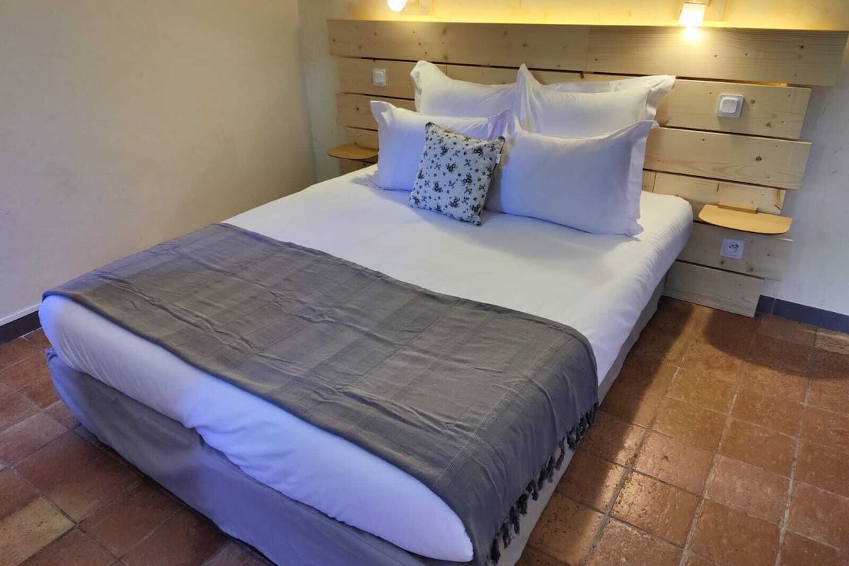 Cama bem arrumada com roupa de cama branca e cinza, cabeceira de madeira e mesas de cabeceira com abajures.