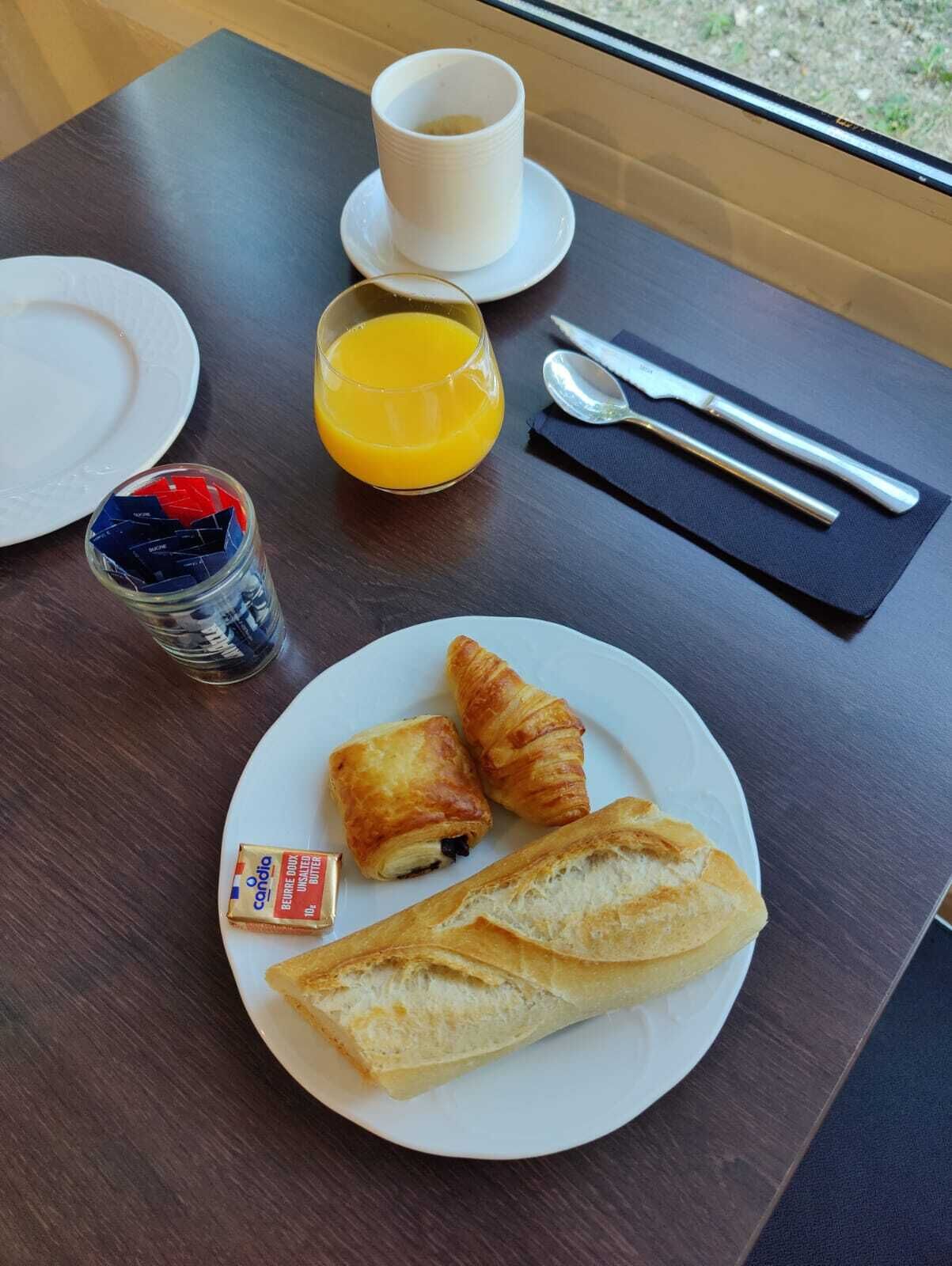 A breakfast setting with croissants, a baguette, a glass of orange juice, a cup, and a small jar of condiments.