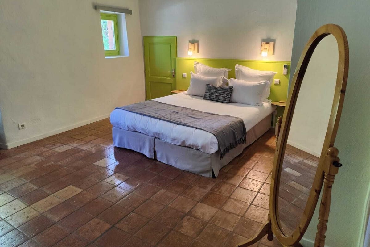 Um quarto com uma cama, um espelho, uma porta verde e uma janela pequena.