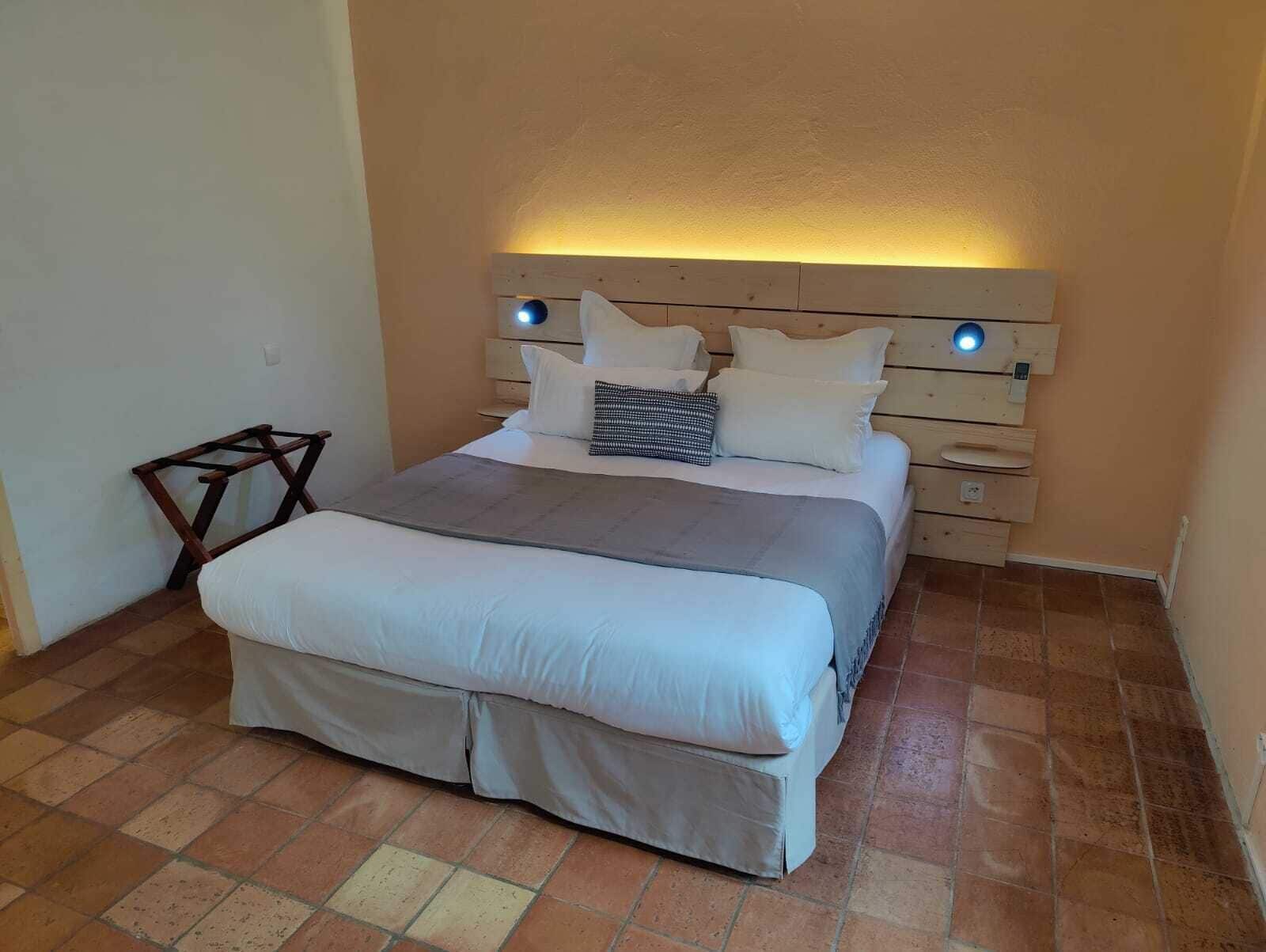 A neatly made bed with a headboard that has built-in lighting, a small wooden stool, and a tiled floor.