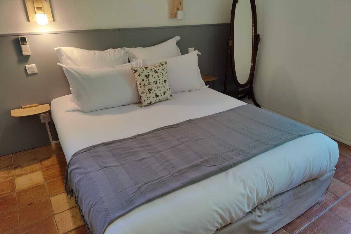 Uma cama bem arrumada com roupa de cama branca e cinza, uma cabeceira de madeira e uma mesa de cabeceira com uma luminária.