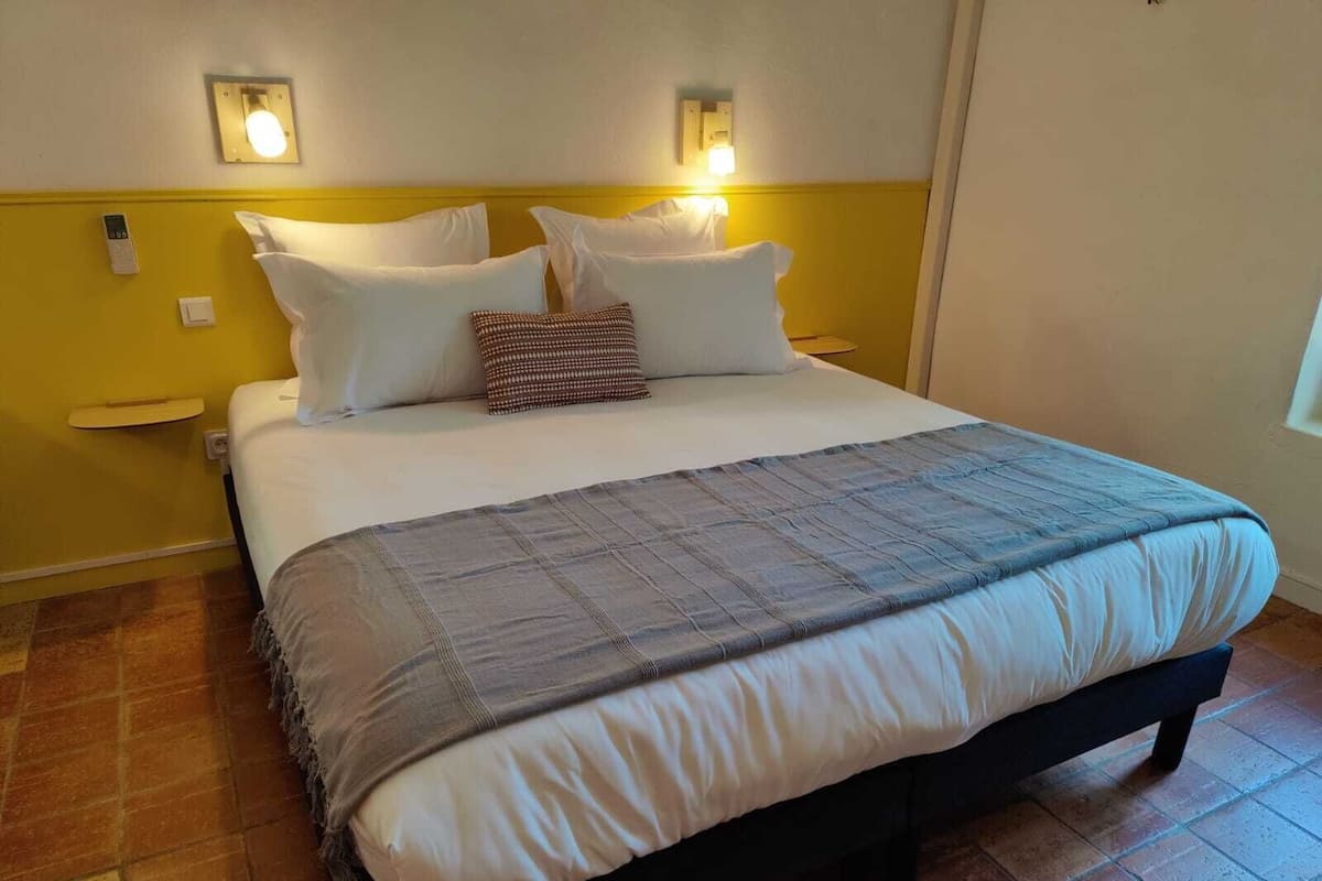 Quarto de hotel com cama, mesa de cabeceira, luminária na parede e tomada.