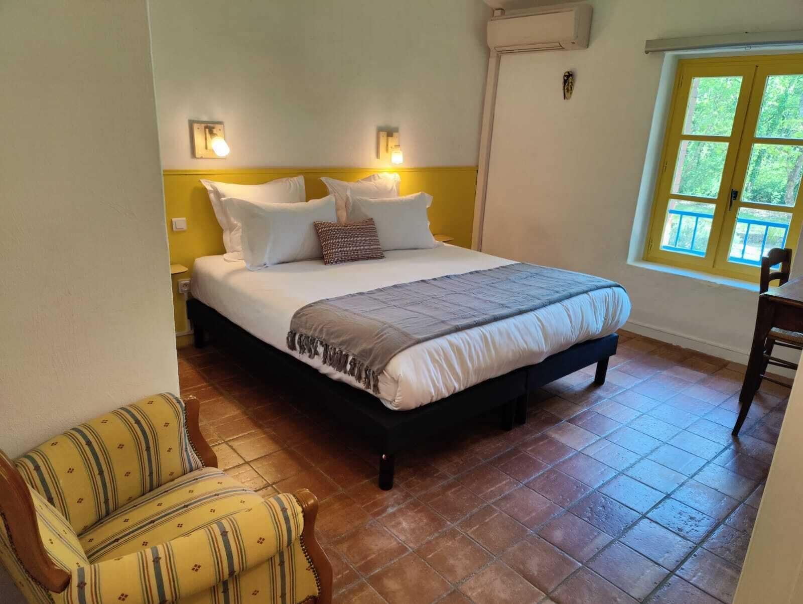 Un dormitorio con cama, mesitas de noche, una silla y ventana con vista a la vegetación.