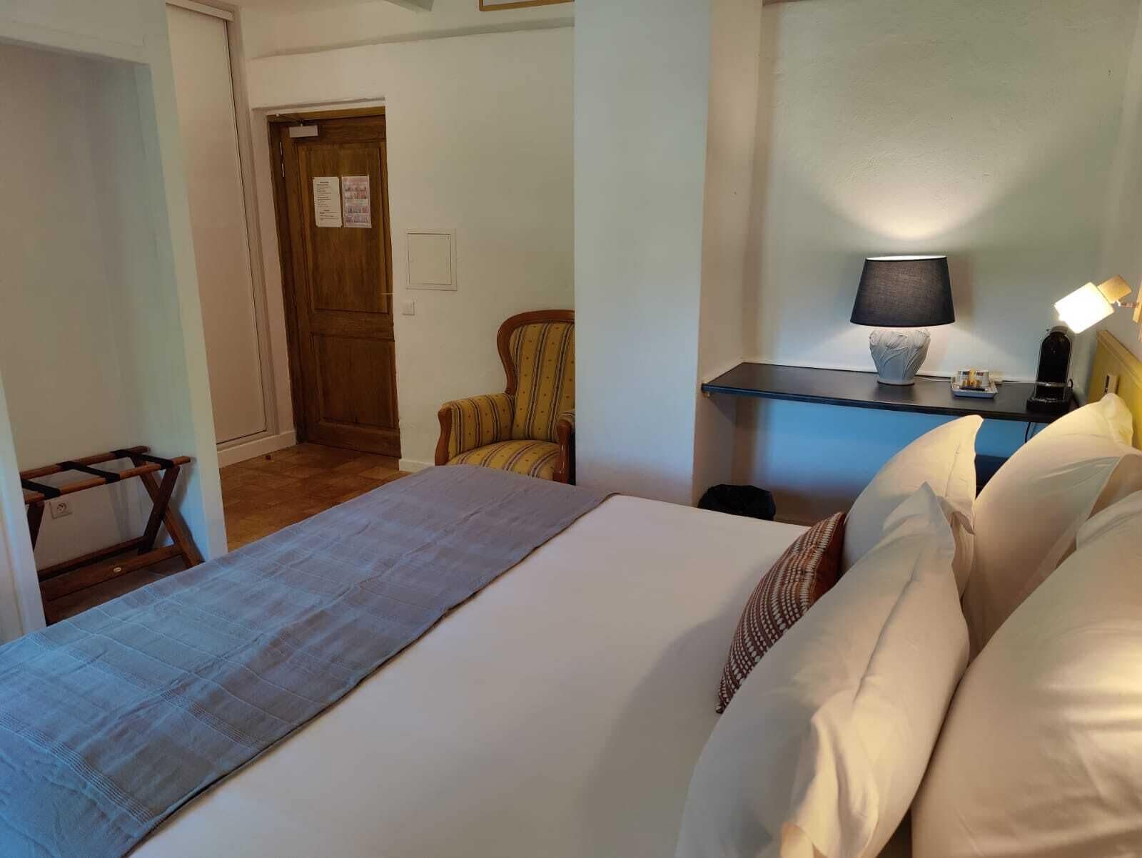 A hotel room with a bed, a chair, a desk with a lamp, and a door.