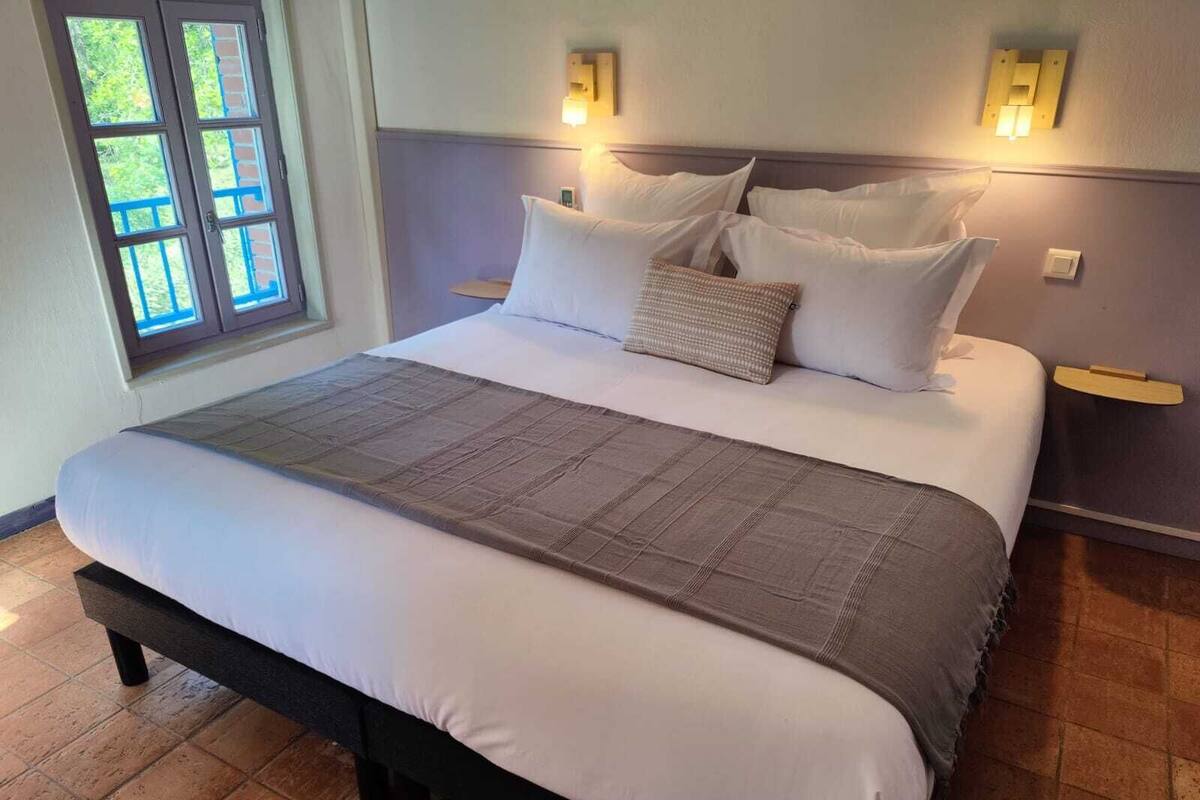 Cama bem arrumada com colcha cinza e almofadas brancas, cabeceira de madeira e janela com vista para o verde.