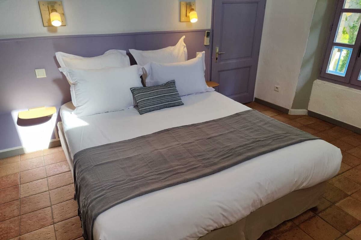 Cama bem arrumada com lençóis brancos e manta cinza, duas luminárias de cabeceira e uma porta com olho mágico.