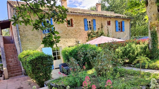 Una casa de piedra con contraventanas azules, un jardín con diversas plantas y flores, y una escalera que lleva a la entrada.
