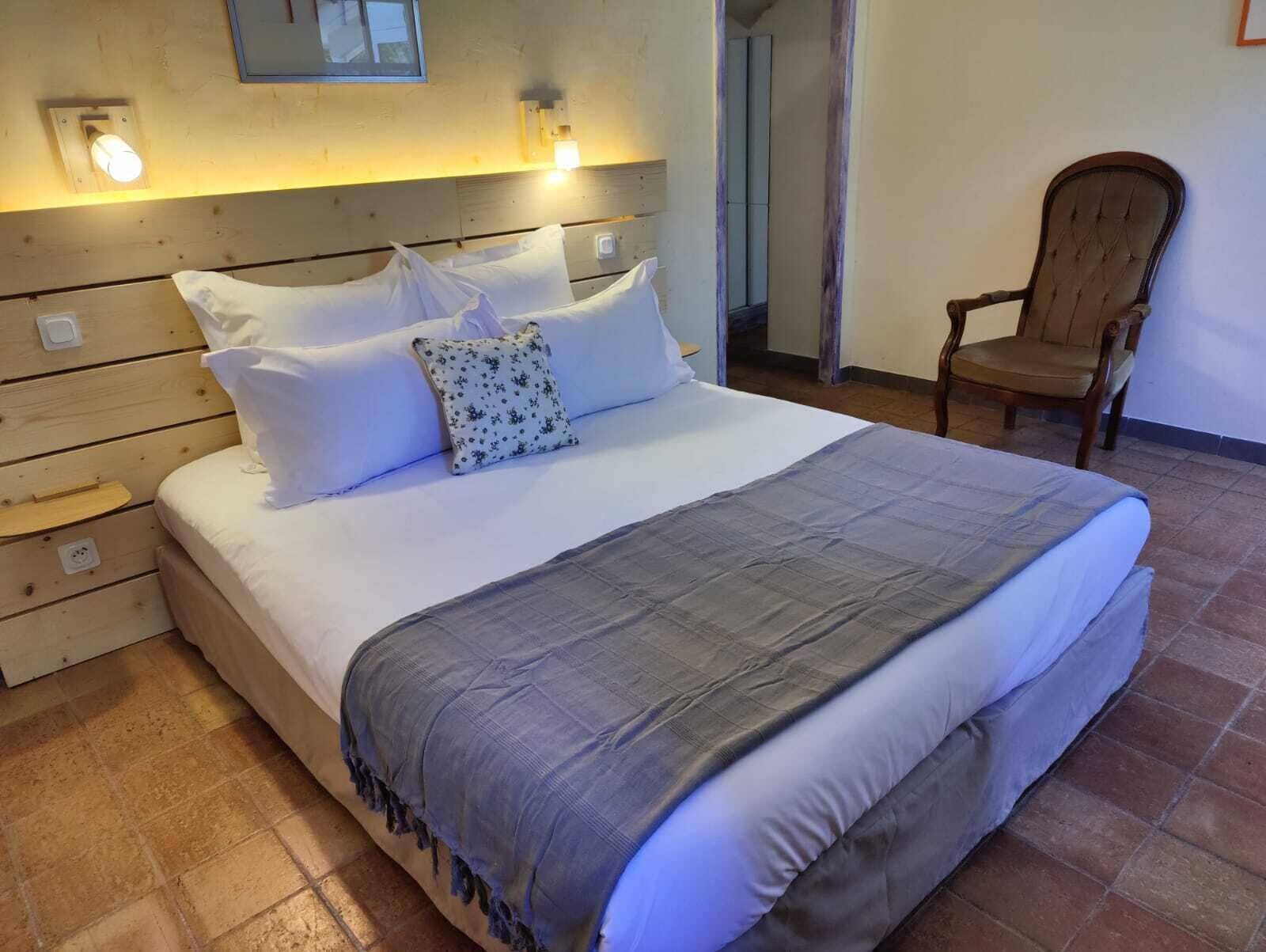 Ein ordentlich bezogenes Bett mit grauer Decke und weißen Kissen, ein Holzkopfbrett und ein Stuhl in einem Zimmer mit gefliestem Boden.