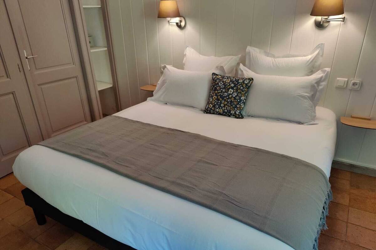 Cama bem arrumada com manta cinza, almofadas brancas e uma almofada floral. Há duas luminárias fixadas na parede, de cada lado da cama.