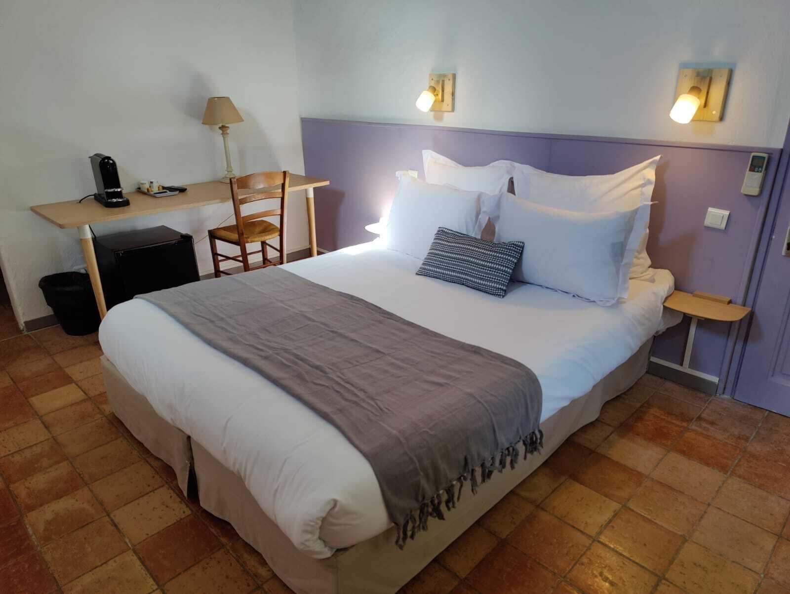 Un lit bien fait, recouvert d’une couverture grise, une table de chevet avec une lampe et une chaise.