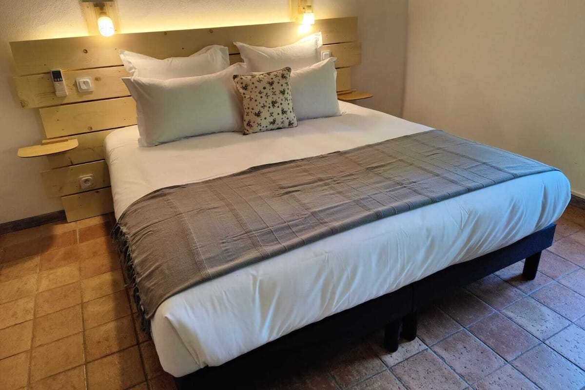 Cama bem arrumada com colcha quadriculada cinza e branca, travesseiros brancos e cabeceira de madeira com luminárias fixadas na parede.