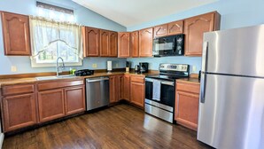 A kitchen with wooden cabinets, stainless steel appliances, and a window with a curtain. - Country Mama's Modern Artsy Cottage  (Sedgwick)