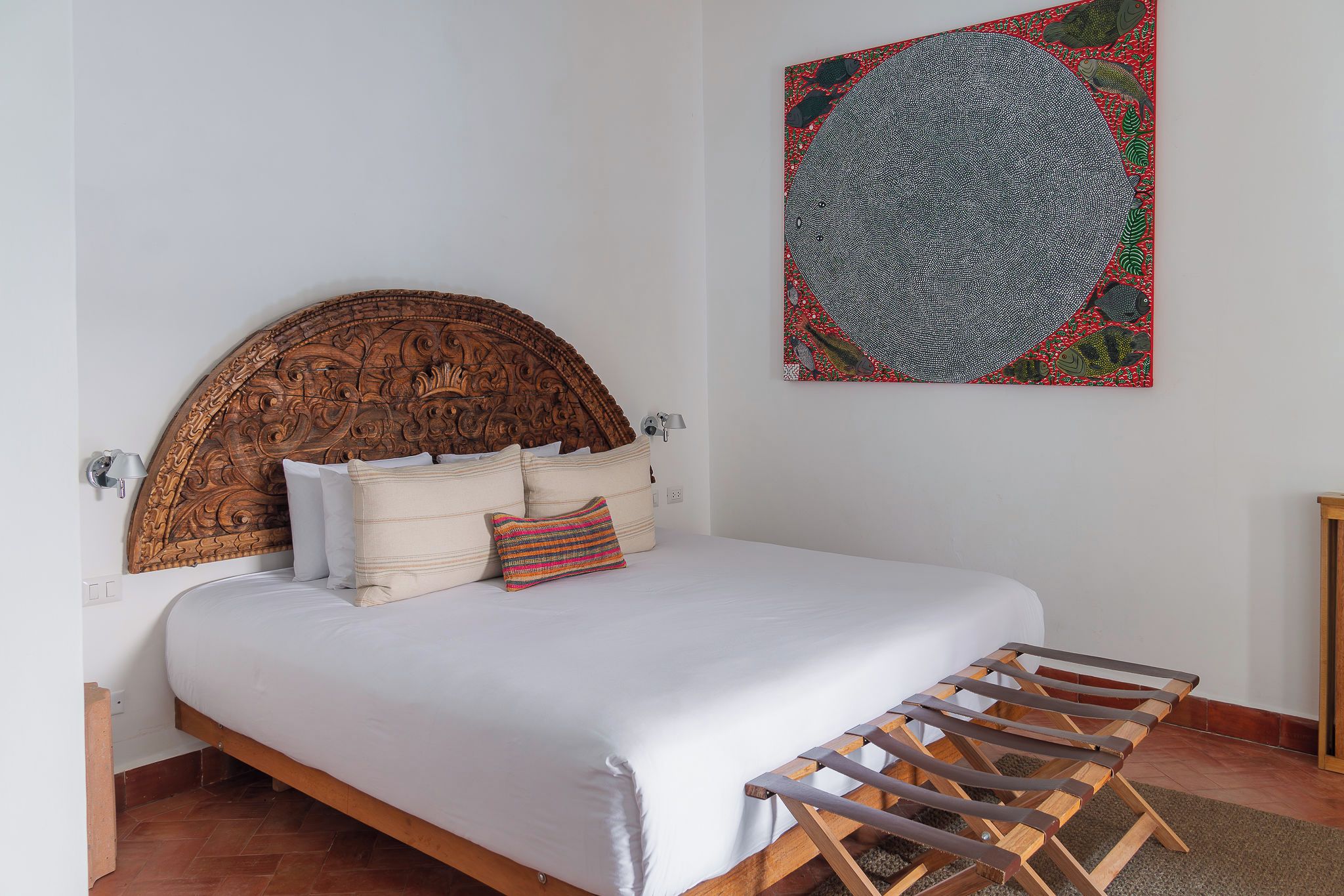 Un dormitorio con cabecera de madera, cama con ropa de cama blanca, almohada colorida, una obra de arte colgada en la pared y un banco de madera.