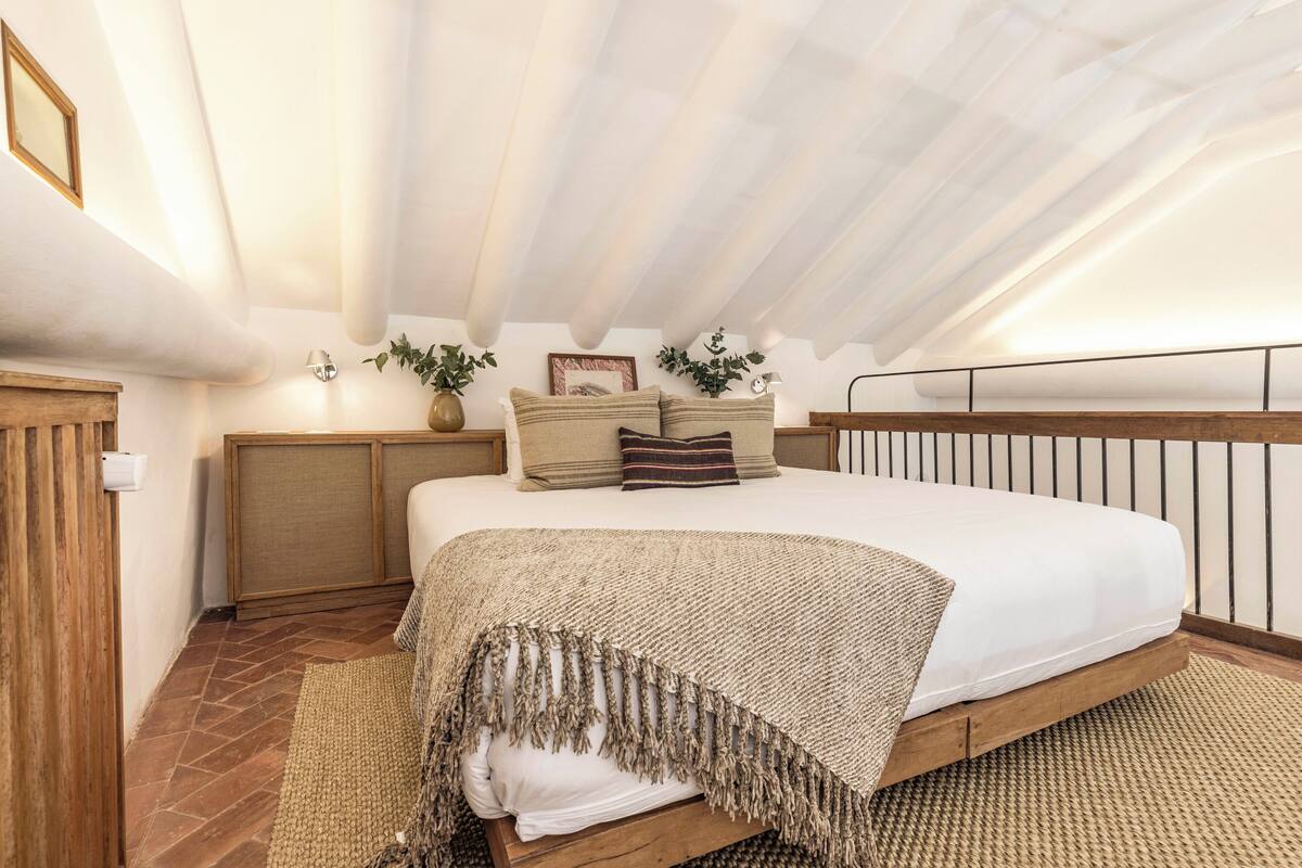Un dormitorio con techo inclinado, una cama con estructura de madera, una manta tejida y una mesita de noche con lámpara.