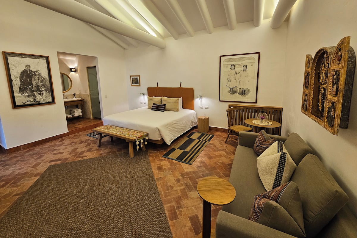 Una habitación de hotel con cama, sofá, una mesita y un banco. Hay cuadros enmarcados en las paredes y un espejo sobre la cama.