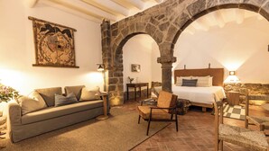 A cozy room with a stone archway, a bed with a wooden headboard, a sofa, a small table, and a chair. - Casa Clara by Ananay Hotels (Cusco)