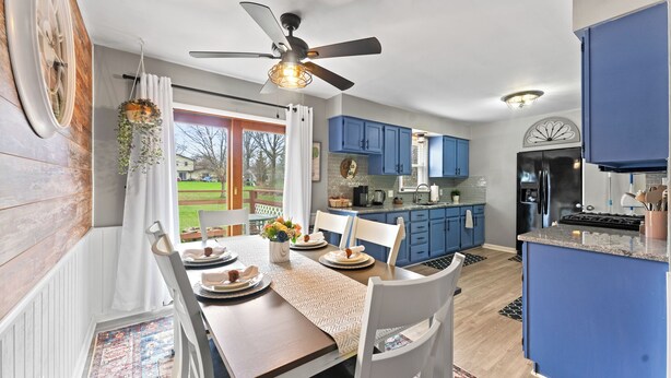A modern kitchen with blue cabinets, a dining area with a wooden table and white chairs, and a ceiling fan.