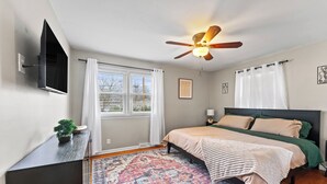 A bedroom with a bed, a ceiling fan, a TV mounted on the wall, and a window with curtains.