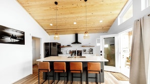 Una cocina moderna con una isla central, techo de madera, luces colgantes y un ventanal grande.