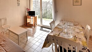A dining area with a table set for four, a television on a stand, and a view of trees through a glass door.