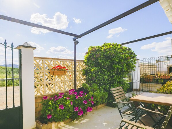 A covered patio with a table and chairs, surrounded by greenery and flowers.