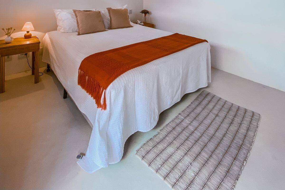 A bedroom with a wooden ceiling, a bed with white bedding and an orange throw, two bedside lamps, and a small table with a lamp.