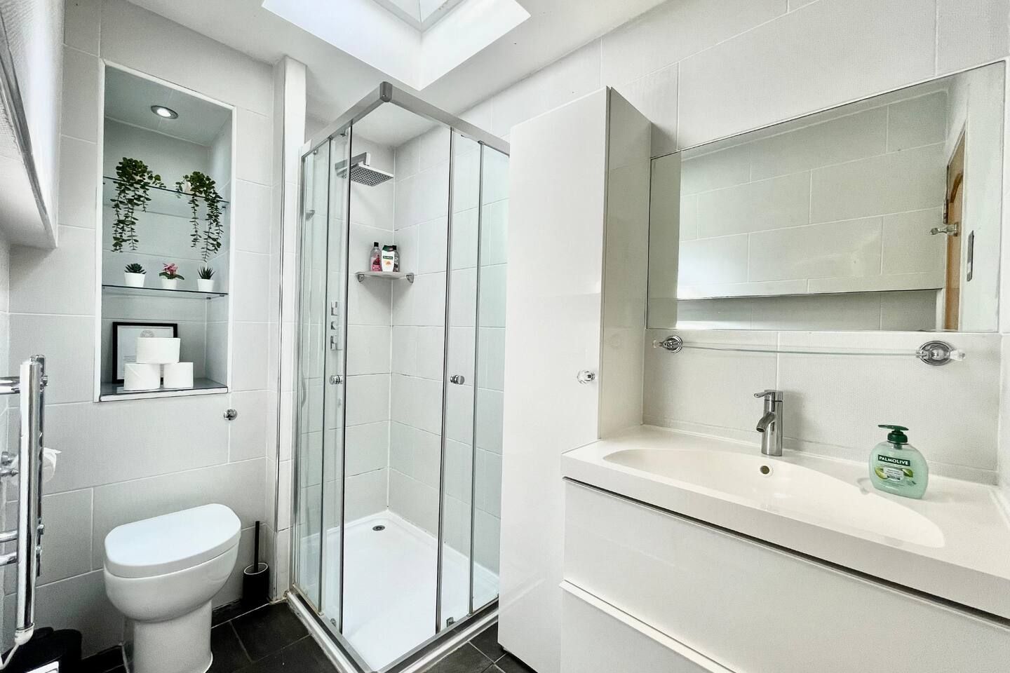 A modern bathroom with a glass-enclosed shower, a toilet, a sink with a mirror, and a shelf with toiletries.
