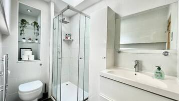 A modern bathroom with a glass-enclosed shower, a toilet, a sink with a mirror, and a shelf with toiletries.