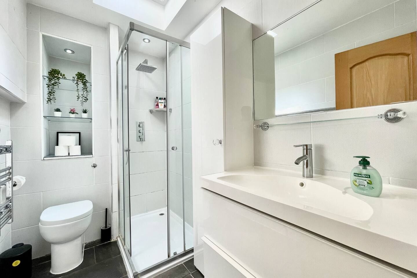 A modern bathroom with a glass-enclosed shower, a toilet, a sink with a mirror, and a shelf with toiletries.