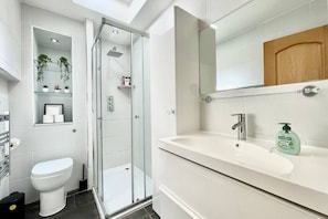 A modern bathroom with a glass-enclosed shower, a toilet, a sink with a mirror, and a shelf with toiletries.