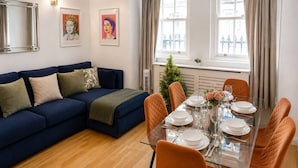 Una sala moderna con un sofá azul, una mesa de comedor de vidrio preparada para cuatro personas y una pequeña planta en maceta.