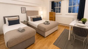Habitación de hotel con dos camas, una mesita con una silla, un radiador y una ventana con cortinas.