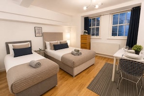 A hotel room with two beds, a small table with a chair, a radiator, and a window with curtains. - 3bdr House + Parking | paddington & little venice (Greater London)