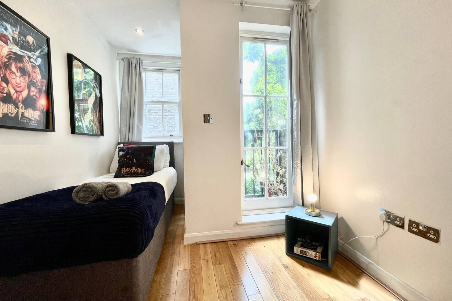 A bedroom with a bed, a window with curtains, a wall-mounted shelf, and a wall socket.