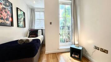 A bedroom with a bed, a window with curtains, a wall-mounted shelf, and a wall socket.