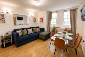 A modern living room with a blue sofa, a dining table set for four, and a flat-screen TV mounted on the wall. - 3bdr House + Parking | paddington & little venice (Greater London)