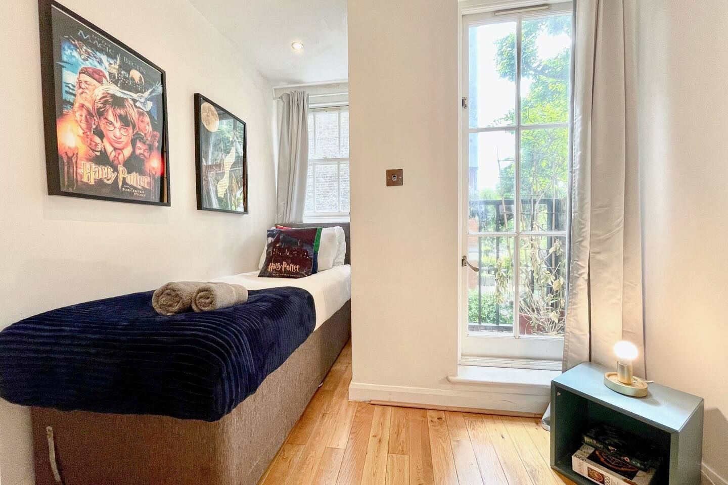 Une chambre à coucher avec un lit, une fenêtre avec des rideaux, un mur orné de tableaux encadrés et une petite table avec une lampe.