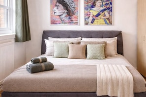 A bed with a grey headboard, multiple pillows, and a blanket. There are two paintings on the wall above the bed.