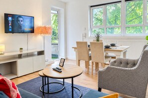 Ein modernes Wohnzimmer mit Fernseher, Essbereich und einem großen Fenster mit Blick auf Grünflächen.
